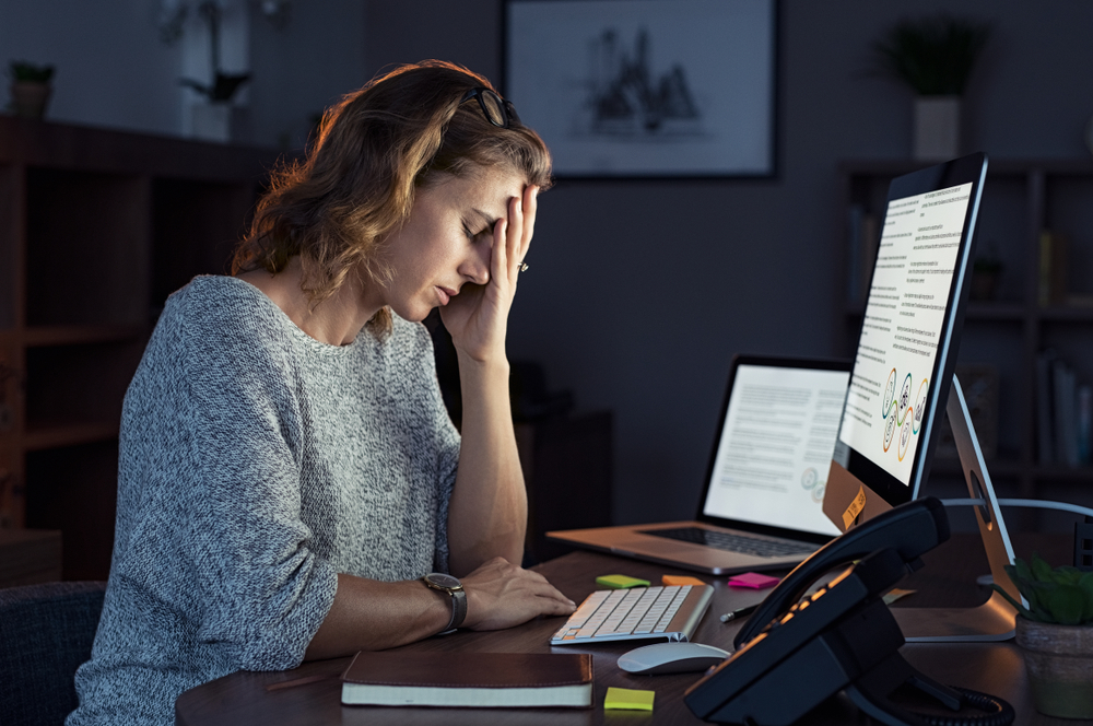 A stressed woman with a hand on her forehead and sitting at a desk in front of a computer